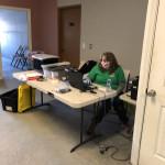 A volunteer sets up and tests the intake station. A volunteer wearing a mask sits at a desk organizing paperwork, equipment, and other items to prepare to take in animals.
